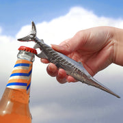 Hand using a shark-shaped bottle opener on a bottle against a cloudy sky.